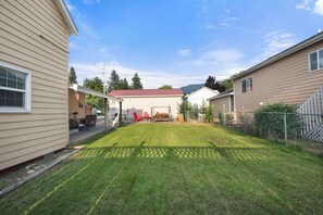 Ein Wohnhausgarten mit Rasen, einer Terrasse und Gartenmöbeln, sowie ein Gebäude mit rotem Dach im Hintergrund.