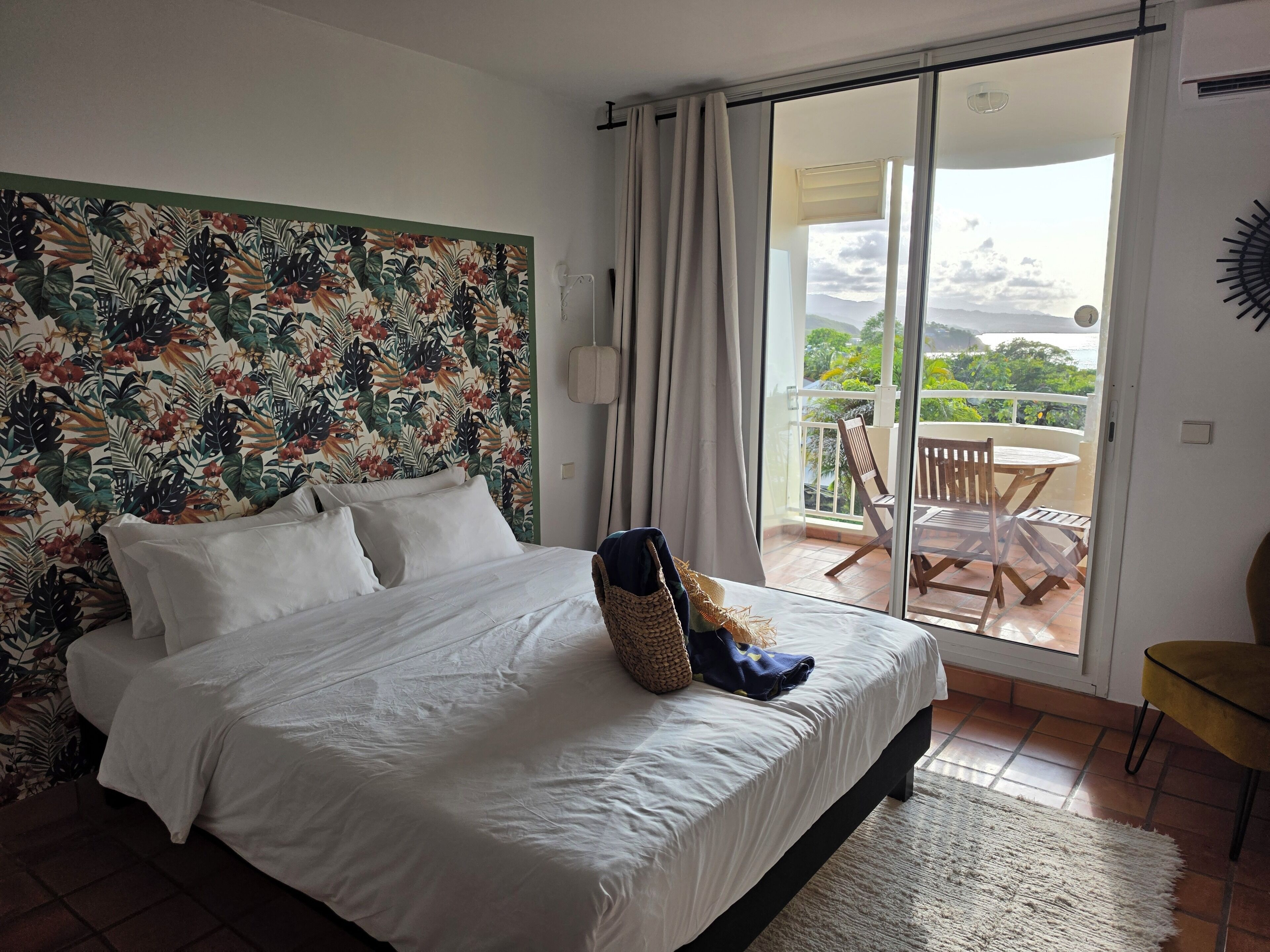 Une chambre dotée d’un grand lit, offrant une vue sur l’extérieur grâce à une porte-fenêtre coulissante, et agrémentée d’une tête de lit sur le thème tropical.