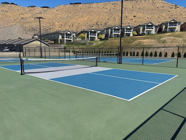 Un court de tennis avec un filet, entouré d’une clôture, et des immeubles d’habitation en arrière-plan.