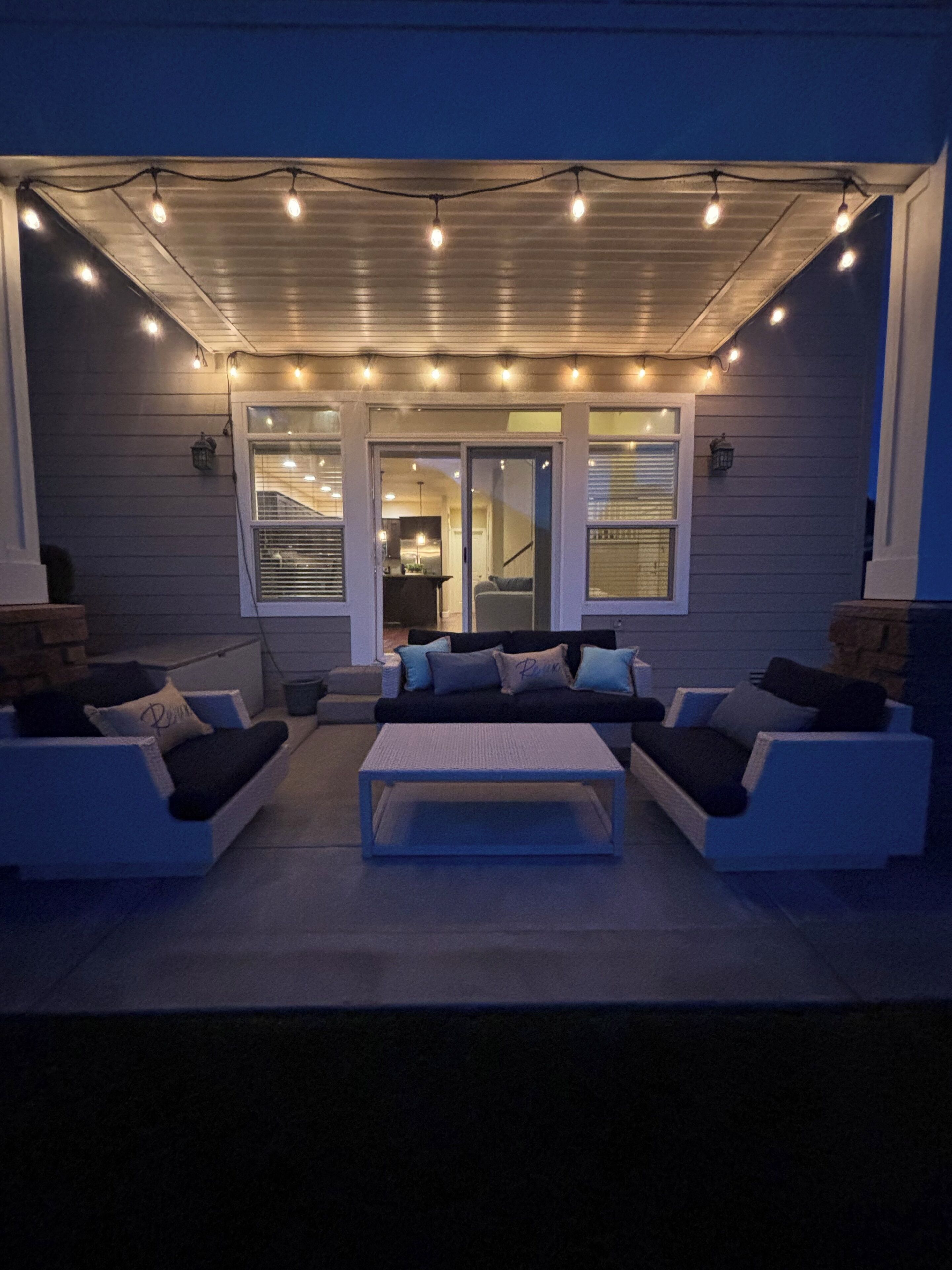 An outdoor seating area with two couches and a coffee table, under a covered patio with string lights.