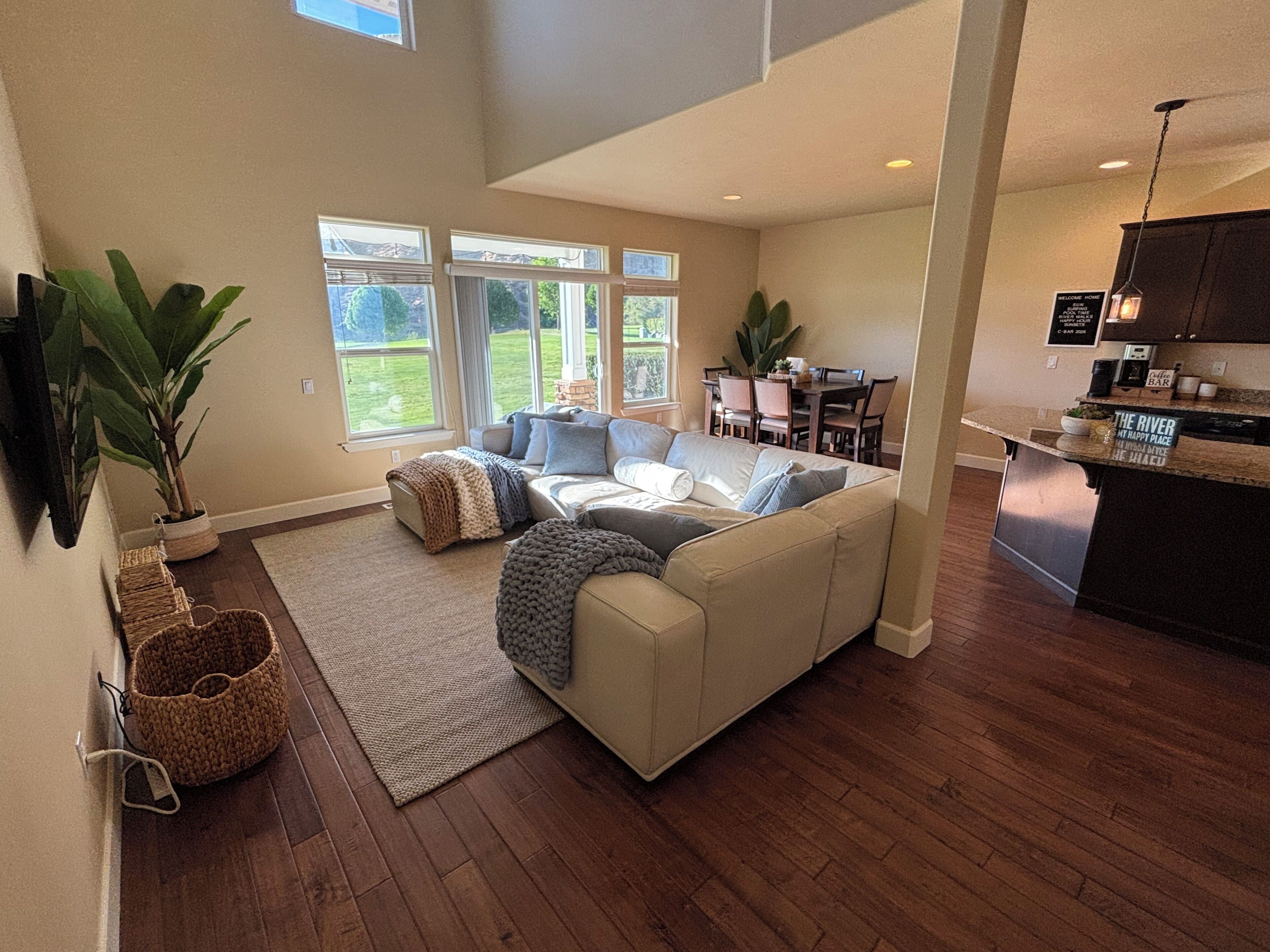 A spacious living area with a large sofa, a coffee table, and a dining table with chairs. There are large windows providing natural light and a view of the outdoors.