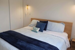 Een net opgemaakt bed met gestreept beddengoed, een donkerblauwe plaid en een kussen met de tekst 'BEACH TIME'.