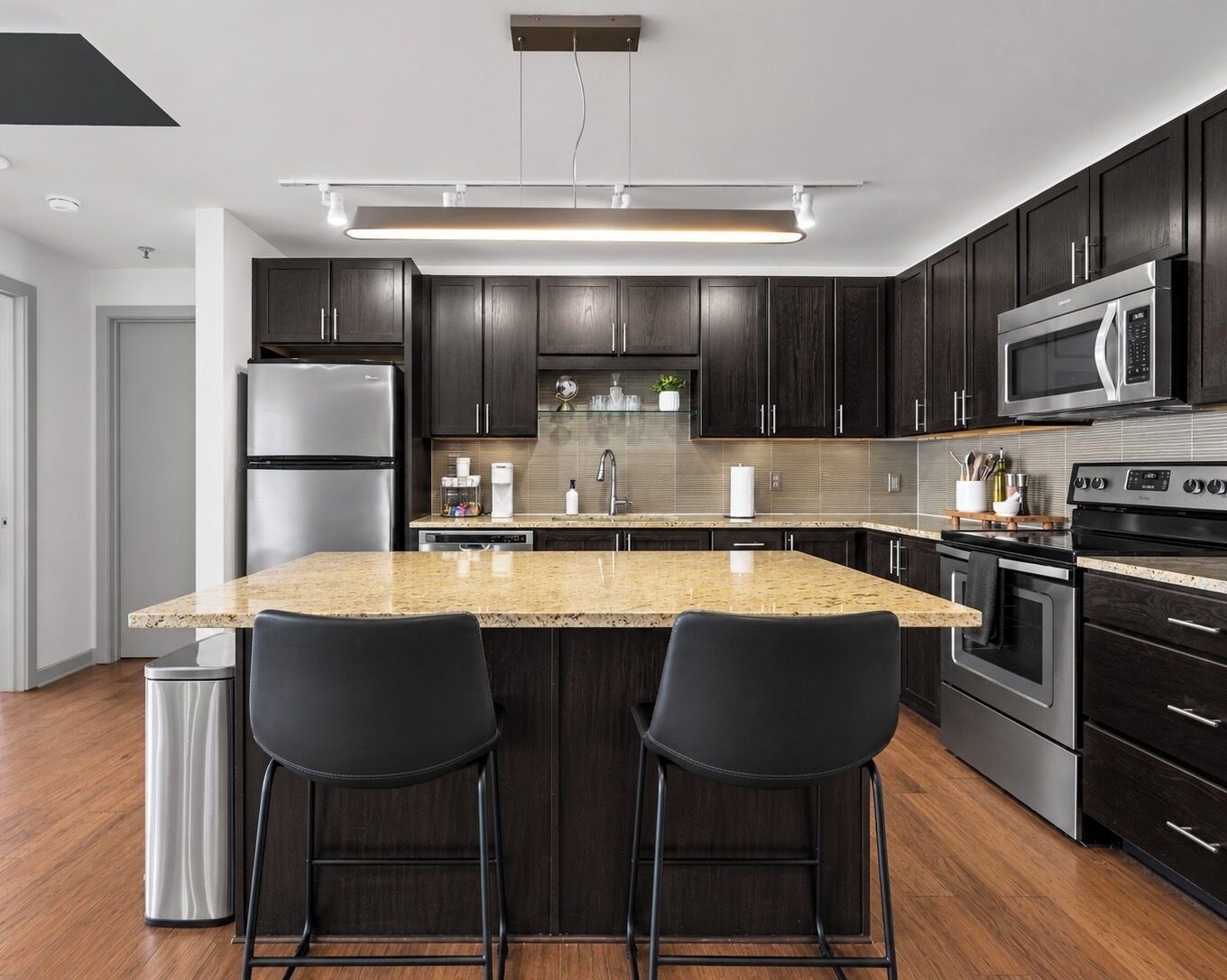 A modern kitchen with dark wood cabinets, stainless steel appliances, a granite countertop, and two black bar stools.