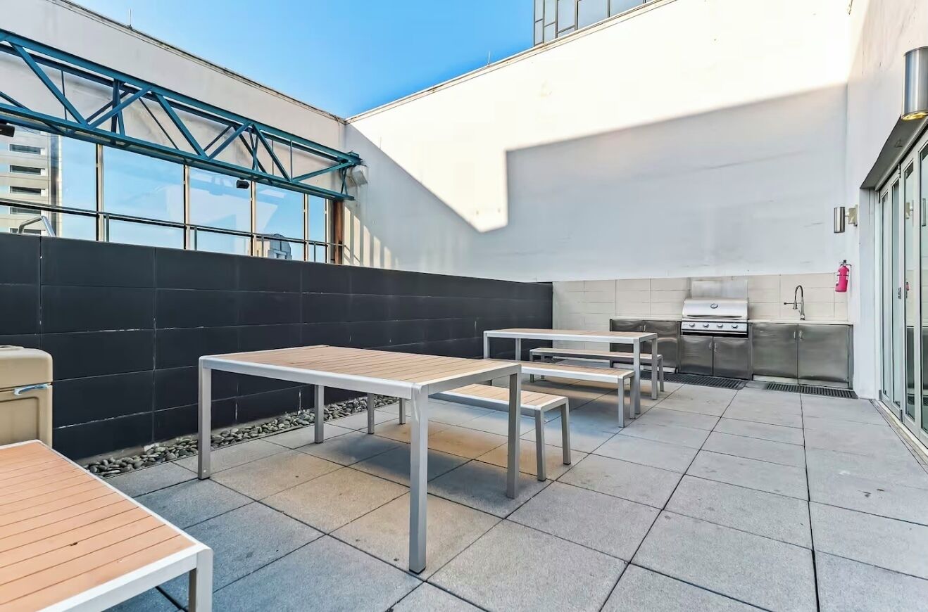 Un patio exterior moderno con mesas y bancos de metal, una parrilla empotrada y una pared negra con estructura de celosía metálica.