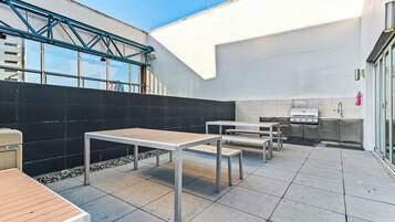 Un patio exterior moderno con mesas y bancos de metal, una parrilla empotrada y una pared negra con estructura de celosía metálica.