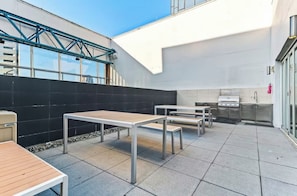 A modern outdoor patio with metal tables and benches, a built-in grill, and a black wall with a metal truss structure. - Luxury Downtown Dallas Penthouse w/ Rooftop Pool, Gym, Free Parking (Dallas)