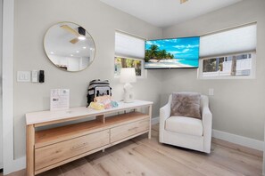 Ein Zimmer mit einem Holzkabinett, einem weißen Sessel, einem runden Spiegel, einem Flachbildfernseher und einem Fenster mit Jalousien.