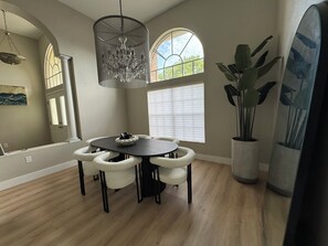 A dining area with a round table, white cushioned chairs, a chandelier, large windows, and a potted plant. - Delightful 3-bedroom house on the golf course (Ocala)