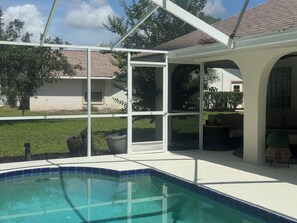 A swimming pool with a clear blue edge, surrounded by a covered patio area with a view of a house and a grassy yard. - Delightful 3-bedroom house on the golf course (Ocala)