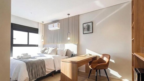 A modern hotel room with a bed, desk, and chair, a window, and a wall-mounted air conditioner. - Studio in Building w/ gym in Jardins Hmj0301 (São Paulo)