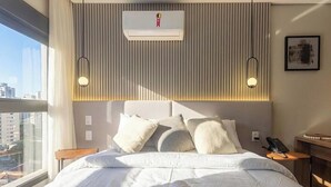 A hotel room with a bed, pillows, an air conditioning unit, a bedside table with a phone, and a window with a view of buildings. - Studio in Building w/ gym in Jardins Hmj0301 (São Paulo)