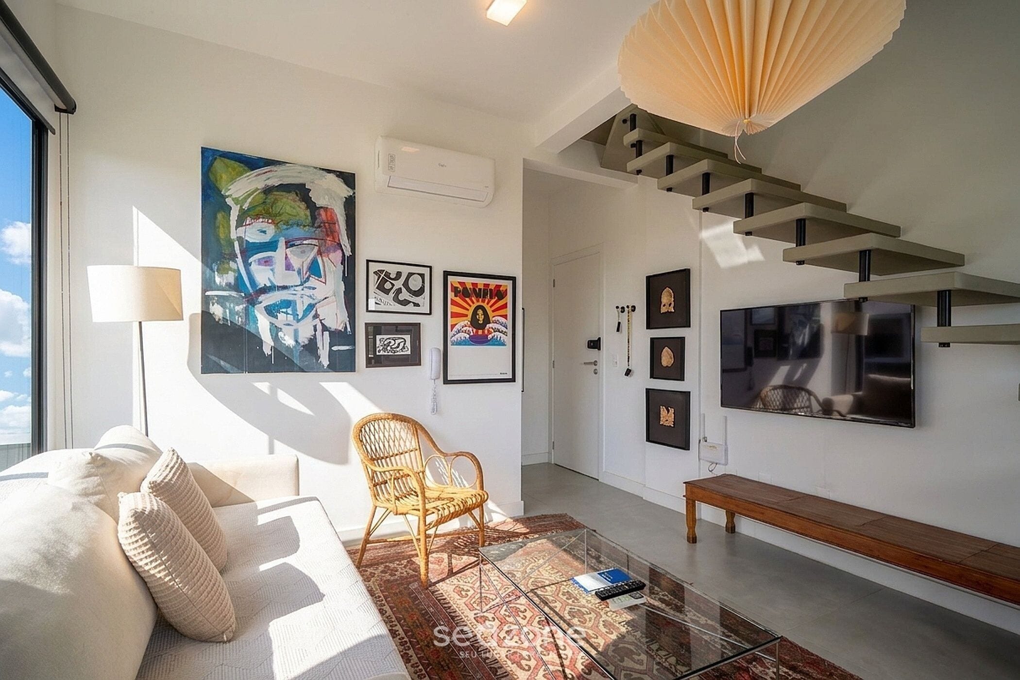 Un salon moderne comprenant un canapé, un fauteuil, un banc et un grand téléviseur. Plusieurs œuvres d’art encadrées ornent les murs, et un escalier au design original se trouve dans la pièce.