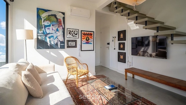 Ein modernes Wohnzimmer mit einer Couch, einem Sessel, einer Bank und einem großen Fernseher. An den Wänden befinden sich mehrere gerahmte Kunstwerke und eine Treppe mit einem einzigartigen Design.