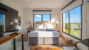 Ein modernes Schlafzimmer mit einem großen Bett, einem Schreibtisch und einem Fernseher. Zwei Fenster bieten Blick ins Freie.
