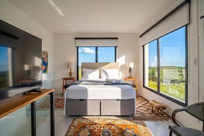 Ein modernes Schlafzimmer mit einem großen Bett, einem Schreibtisch und einem Fernseher. Zwei Fenster bieten Blick ins Freie.