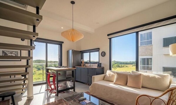 Um interior moderno com sofá, mesa de jantar e cozinha, destacando grandes janelas e uma escada.