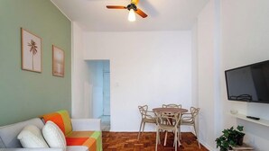 Un soggiorno con divano, tavolo da pranzo con sedie, ventilatore a soffitto, televisore e una porta che dà su un'altra stanza.