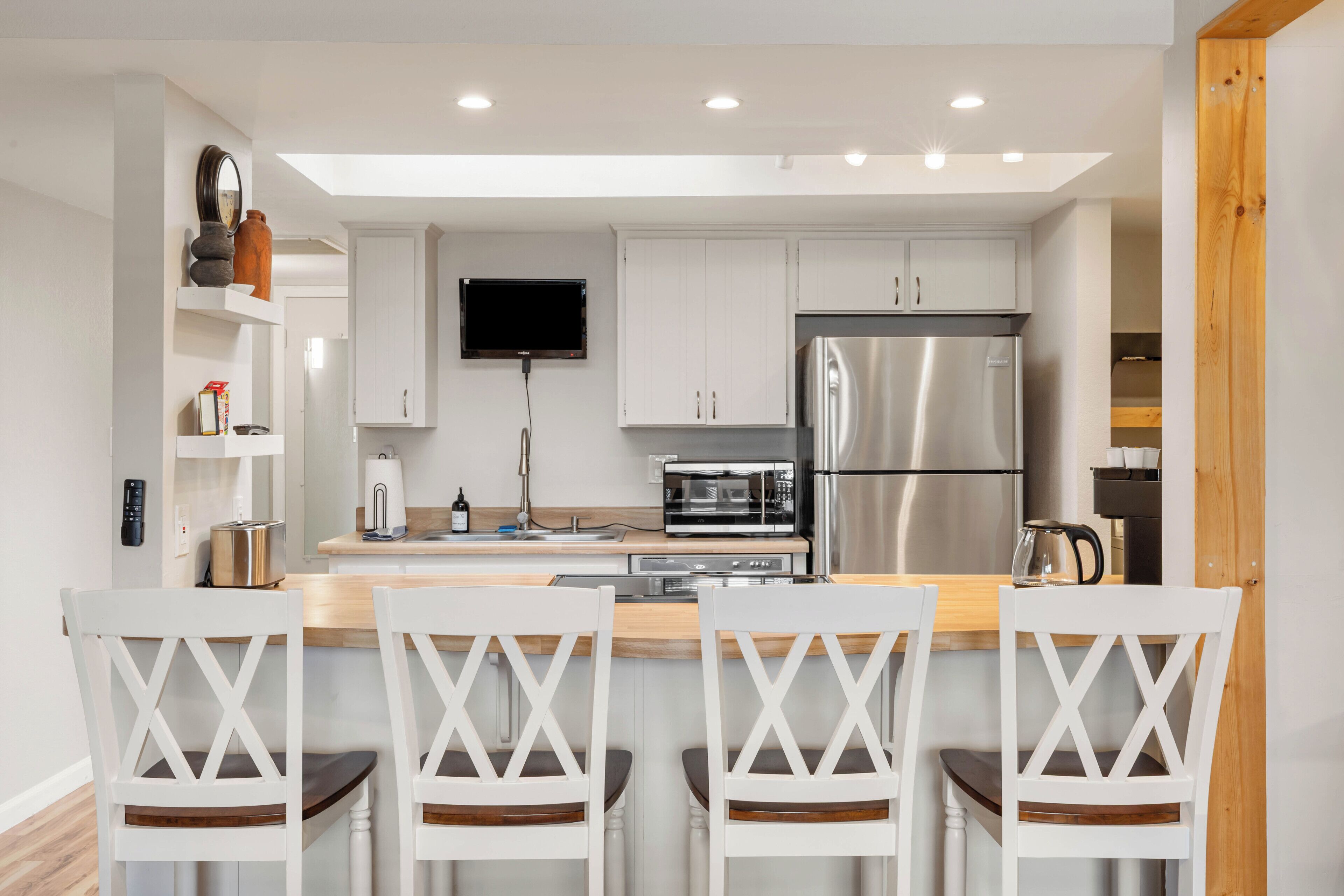A modern kitchen with white cabinetry, a stainless steel refrigerator, a microwave, and a flat-screen TV. There are four white chairs with wooden seats and backs.