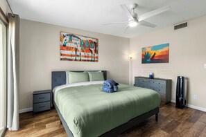 Une chambre à coucher comprenant un lit, une table de chevet, une commode, un ventilateur de plafond et deux tableaux accrochés au mur.