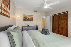 Une chambre à coucher comprenant un lit, une table de chevet, une lampe, un tableau, une armoire et un ventilateur de plafond.