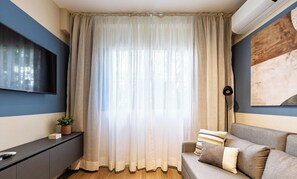 Un saló amb un finestral gran cobert per cortines lleugeres, un sofà gris amb coixins estampats, una taula petita amb una planta i un televisor pla penjat a la paret.