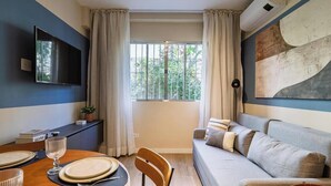 Un soggiorno moderno con un divano, un tavolo da pranzo apparecchiato con piatti e un bicchiere, una TV a parete e una finestra con tende.