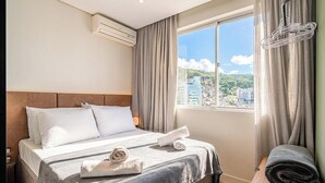A hotel room with a bed, a window with curtains, an air conditioning unit, and a view of a cityscape. - Studio in Building w/ View Downtown Eva0706 (Florianópolis)