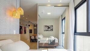 A modern hotel room with a bed, a desk, and a view of the city through large windows. - Studio in Condo w/ Pool Downtown Itapema Ffs0606 (Itapema)