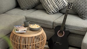 Eine schwarze Gitarre, eine Popcornschale, ein Buch und eine Tasse auf einem runden, gewebten Tisch in einem Wohnzimmer mit grauen Sofas und gemusterten Kissen.