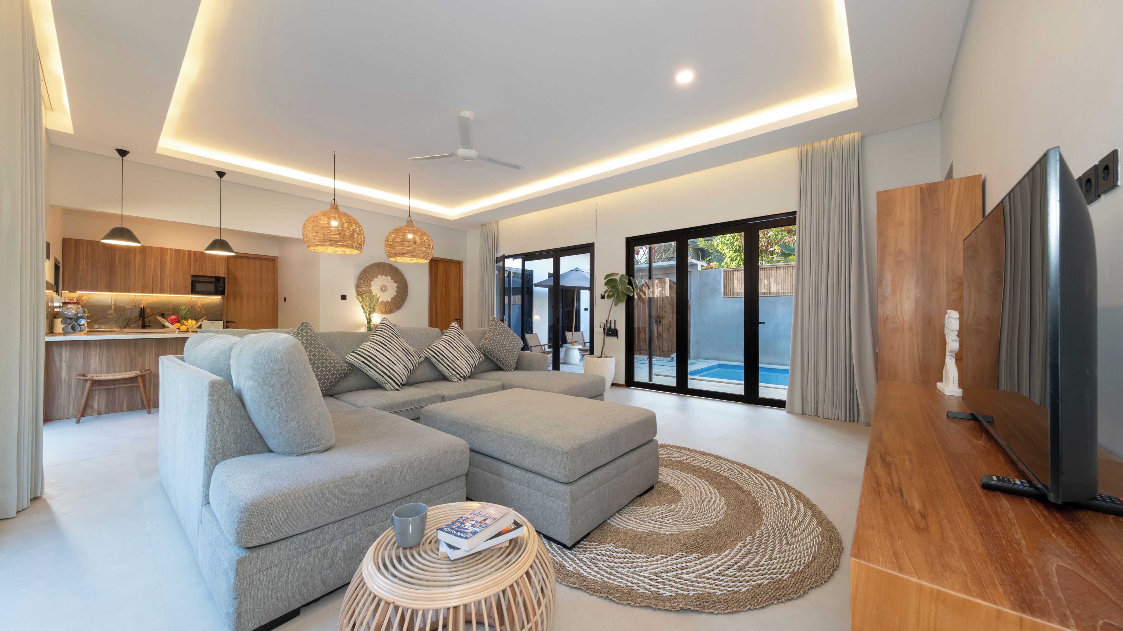 Un salon moderne avec un canapé d’angle, un coin repas avec une cuisine en arrière-plan et une porte-fenêtre coulissante donnant sur une piscine.