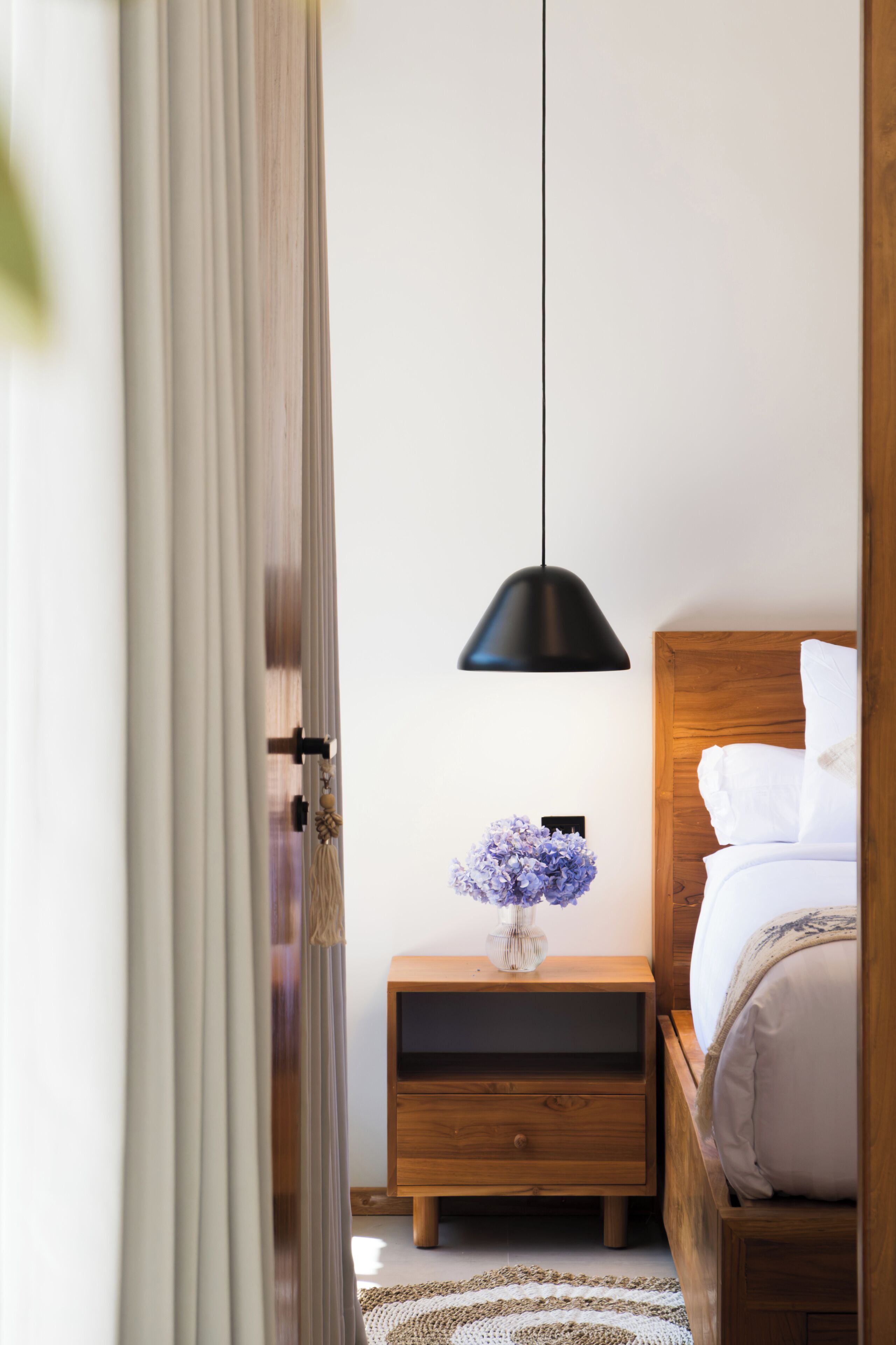 Un dormitorio con cabecera de madera, una mesita de noche con un florero, una lámpara colgante negra y una mesita auxiliar de madera.