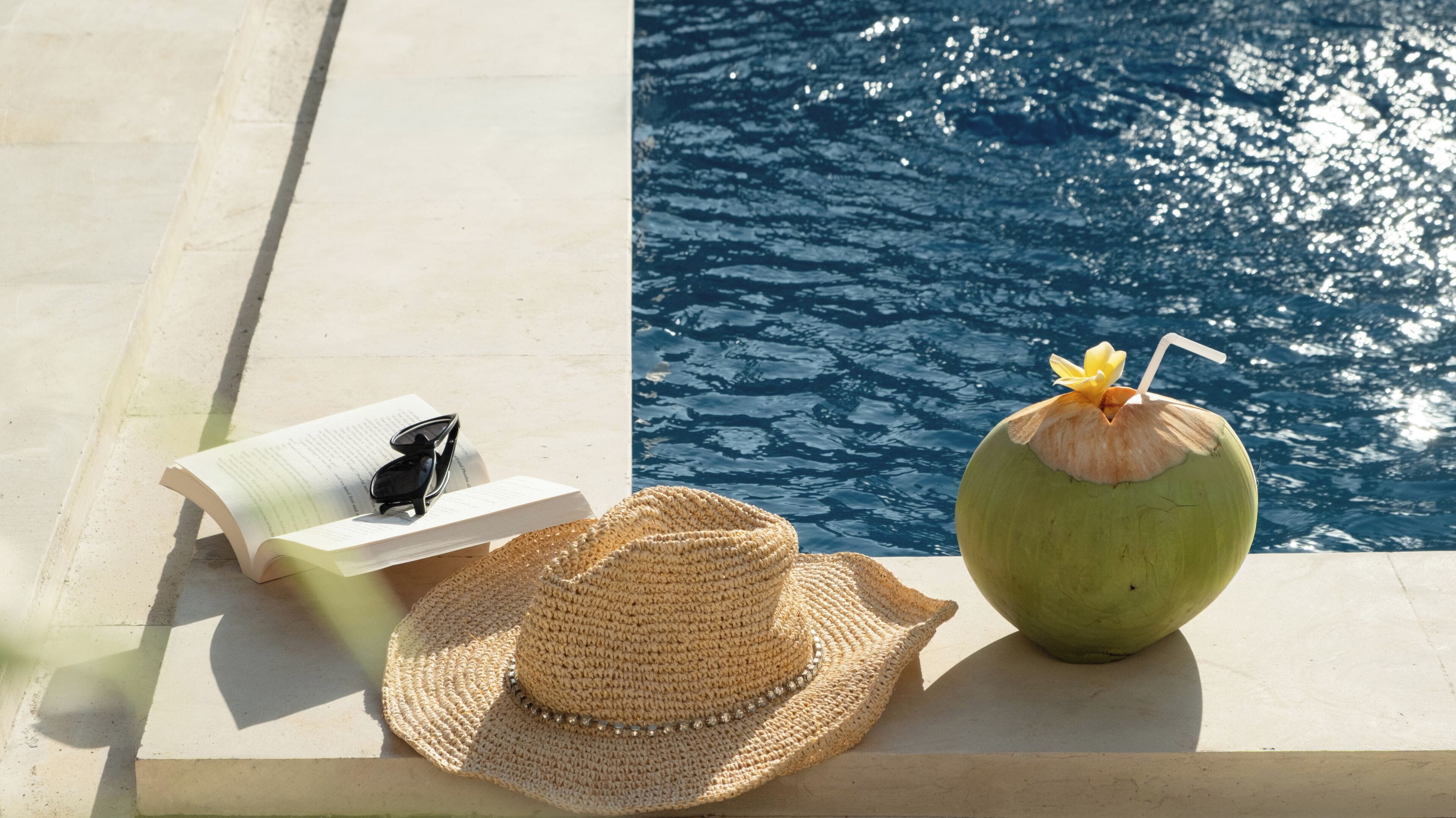 A poolside setting with a straw in a coconut, a book, and a straw hat.
