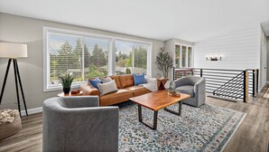 Ein modernes Wohnzimmer mit einer Couch, Sesseln, einem Couchtisch und einem großen Fenster mit Jalousien.
