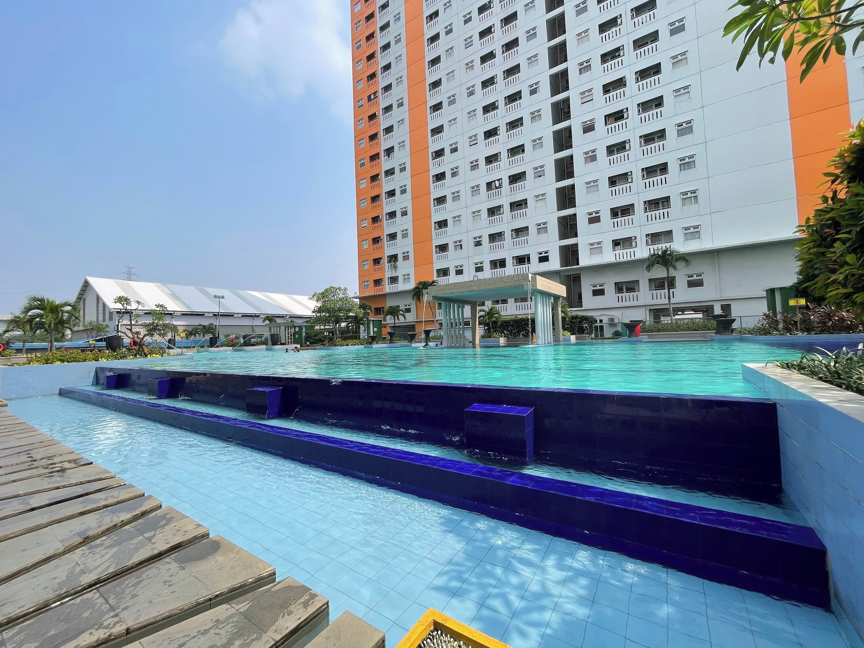 A swimming pool with a wooden deck and steps leading into the water, surrounded by a high-rise building with balconies.
