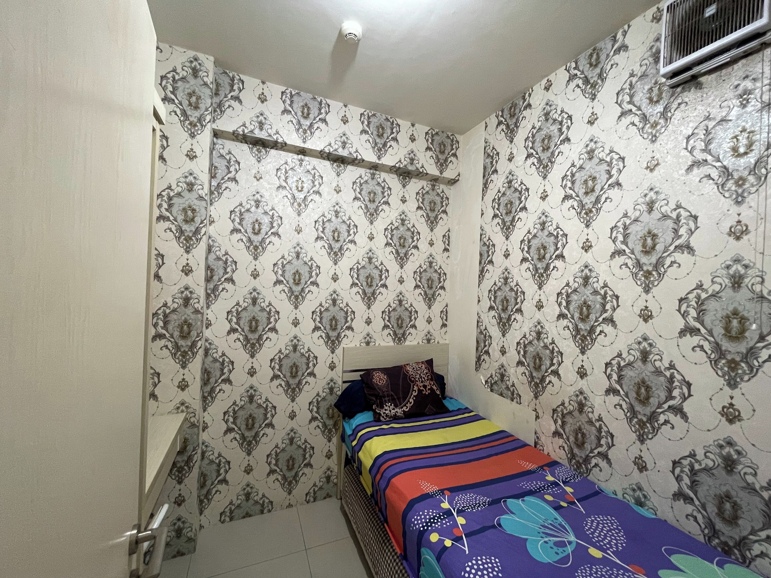 A small, narrow room with patterned wallpaper, a bed with a colorful bedspread, and a window with a vent.