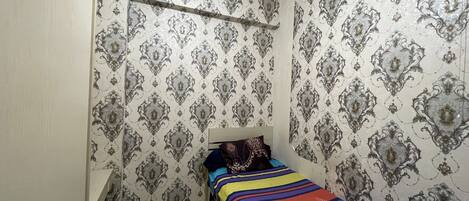 A small, narrow room with patterned wallpaper, a bed with a colorful bedspread, and a window with a vent.