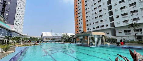 A swimming pool surrounded by high-rise buildings with balconies, a covered area, and some greenery.