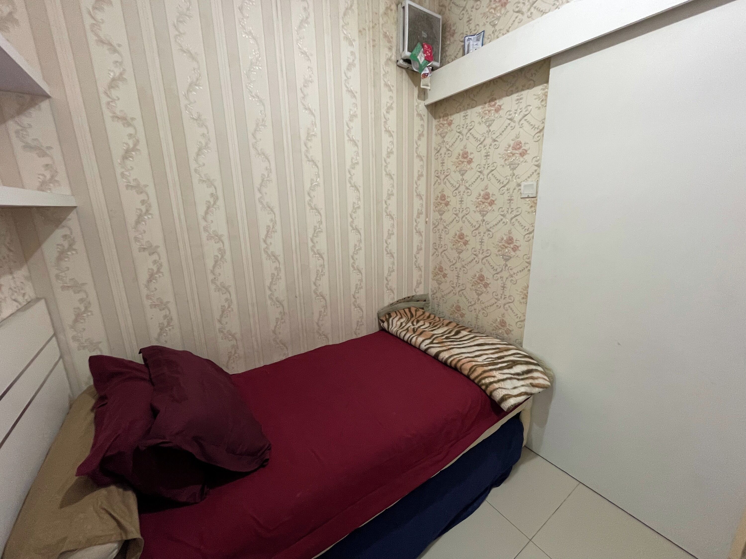 A small room with a bed, a shelf with items, and patterned wallpaper.