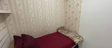 A small room with a bed, a shelf with items, and patterned wallpaper.