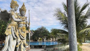 Una estatua de dos figuras con atuendo elaborado, una piscina y palmeras al fondo.