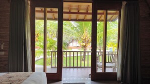 Una habitación con cama, una puerta de madera que da a un balcón y vista a un jardín con palmeras.