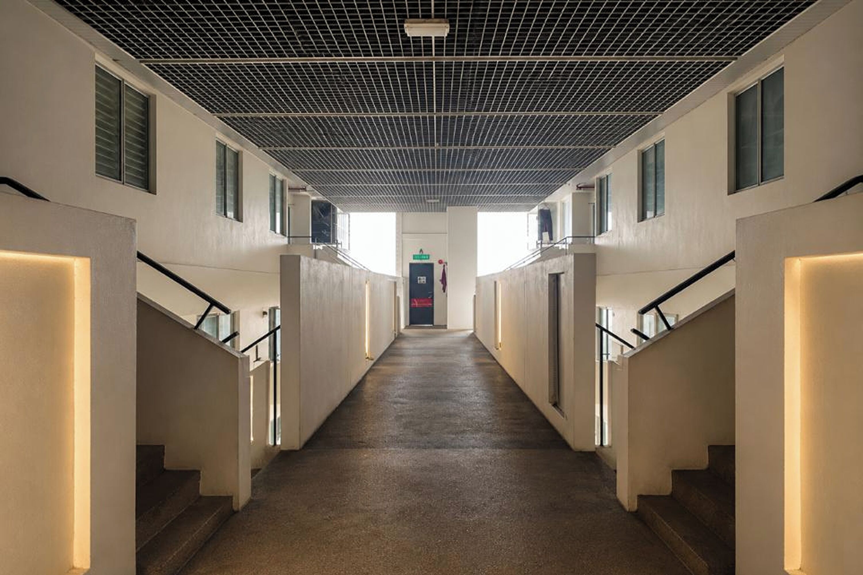 A corridor with a grid ceiling, flanked by walls with windows and doors, leading to a central point.