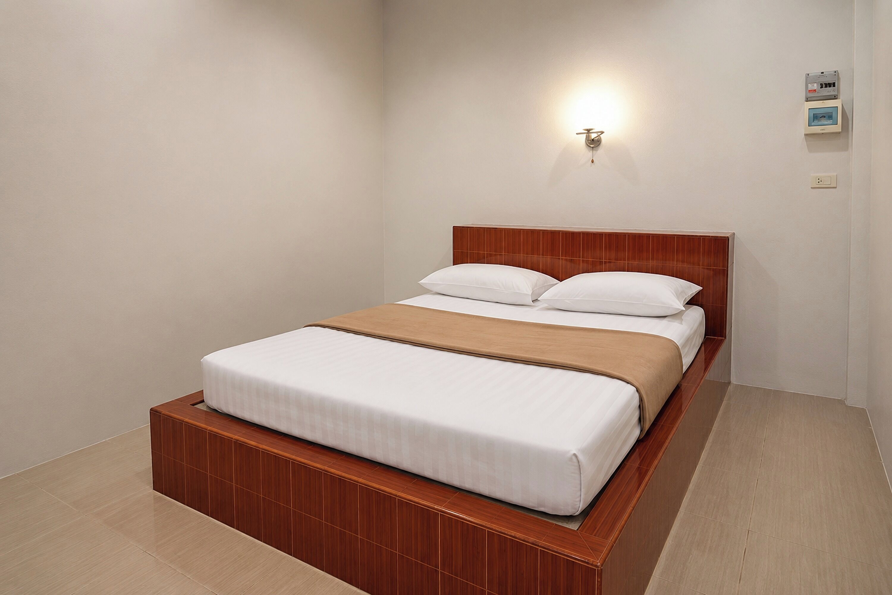 A single bed with a wooden headboard and a beige blanket, a wall-mounted light fixture, and a small picture frame on the wall.