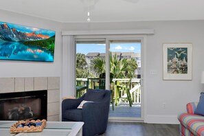 A living room with a fireplace, a large TV, a sofa, and a balcony with outdoor furniture. - Barrier Dunes 451 - 40 Lime Breeze (Port St. Joe)