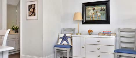 Una habitación con pared blanca, un cuadro enmarco, una lámpara, un cojín con forma de estrella de mar y una estantería con libros.