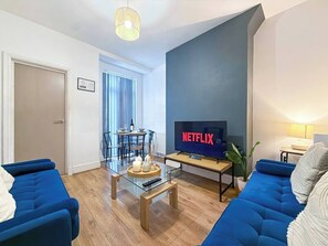 A living room with a blue sofa, a glass coffee table, a flat-screen TV, and a dining area with a table and chairs. - 3 BR House | Sleeps 7 | Free Parking | Fast Wi-Fi (Greater Manchester)