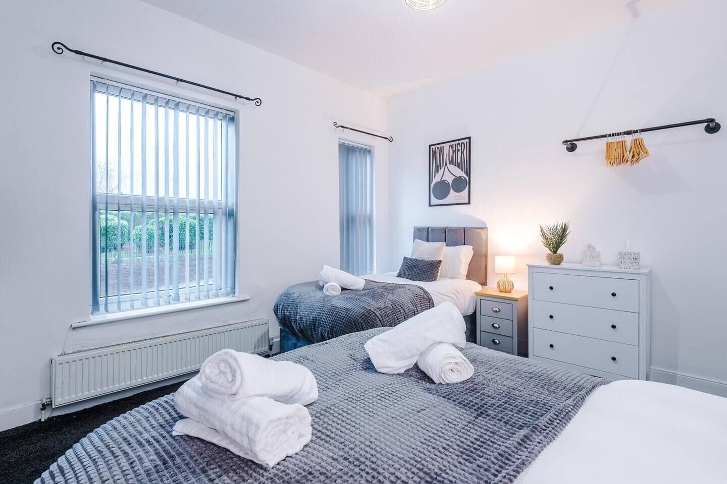 Une chambre d’hôtel avec deux lits, une commode blanche, une table de chevet et une fenêtre avec des stores.