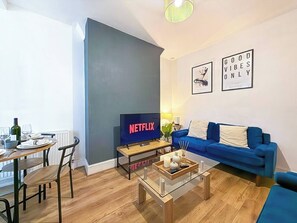 A modern living room with a blue sofa, a coffee table, and a TV displaying 'NETFLIX'. - 3 BR House | Sleeps 7 | Free Parking | Fast Wi-Fi (Greater Manchester)