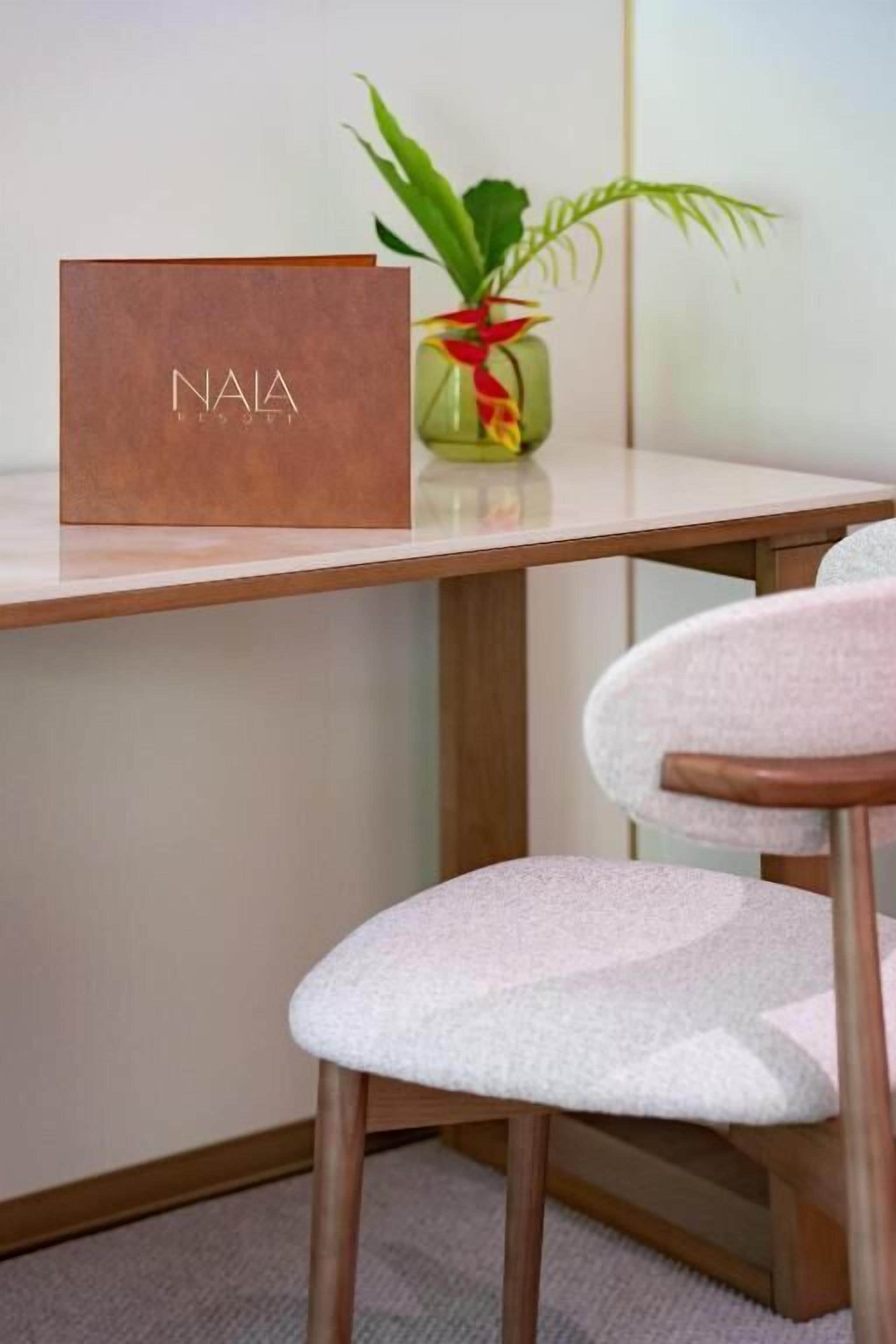 Una silla de madera con un cojín blanco, una caja marrón con el texto 'NAIA' y una planta en un florero de vidrio sobre una mesa.