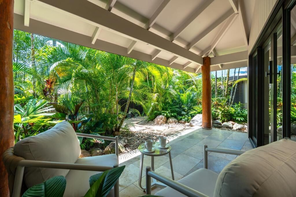 A covered outdoor area with a seating arrangement, a small table, and a view of a tropical garden.